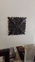 Square metal decorative wall hanging mounted on wall with intricate floral and scroll design in dark finish