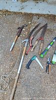 Photo showing yard tools laid on ground including pole saw, large hedge shears, hand saw, pruning saw, and pruning shears