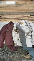 Two coats laid side by side, one maroon leather with quilted pattern and the other white camouflage with gray patches and multiple pockets.