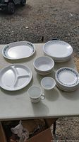 Photo showing oval serving plate, stack of small plates, multiple bowls, partitioned plate, and a cup on a table outdoors.