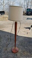 Full view of the mid-century style floor lamp with wooden tapered base and beige fabric drum shade, shown outdoors.