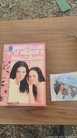 Gilmore Girls DVD box and disc laid out on wooden surface showing box front cover with main characters and one disc out of case.