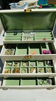 Open white jewelry box showing green felt compartments filled with various costume jewelry pieces including rings, brooches, and pins