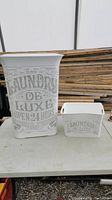 Tall white laundry hamper with vintage-style grey lettering beside smaller white basket with handles and matching grey lettering on a table.