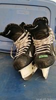 Front and top angled view of pair of black Bauer ice hockey skates, showing laces, tongue, and blade holders.