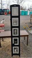 Full view of black photo tower frame standing upright with five 8x10 photo slots, outside in natural light with outdoor background including construction equipment and animal.