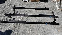 Three black metal adjustable rails for queen bed frame laid out on gravel surface. Some wheels attached, missing two wheels total on pieces.