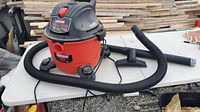 Shop Vac wet/dry vacuum on white table with flexible black hose and cleaning nozzle attached, placed outdoors in front of stacked wood.