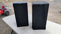 Pair of KEF Q-series speakers with grilles on, positioned outdoors on a white folding table.