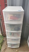 Full view of white plastic 5-drawer storage tower on wheels placed outdoors showing visible contents through drawers.