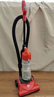 Front view of Dirt Devil upright vacuum cleaner showing red base, clear dust canister, black hose and red handle.