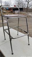 Foldable metal work stool with wooden seat showing wear and damage, taken outdoors on white surface