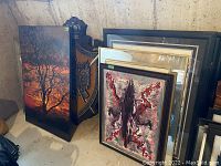 Overview photo showing multiple framed wall art pieces including a photo of a sunset with dark trees, an abstract painting, and a carved wood framed mirror with metallic detail.