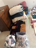Stack of various quilts, blankets, and comforters piled on the floor and leaning against a dresser. Includes a variety of colors and materials.