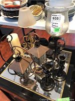 Photo showing multiple candle holders and one white pillar candle on a mirrored surface, including curved brass holders, a small lamp, and black metal hand-shaped holders.