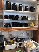 Three shelves showing five black canisters top shelf, twenty-two black mugs and two black teapots on middle shelves, plus bins with cleaning cloths, hand tools, tape, and batteries on bottom shelf