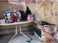 Photo showing a table with assorted vintage Christmas decorations including porcelain Santa, small trees, boxes of lights, and angel figurines, plus a large box on the floor with more decorations.