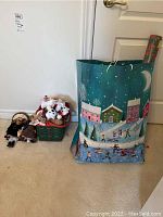 Photo with two porcelain dolls beside a small green basket of plush snowman toys and multiple holiday gift bags including a large blue bag with winter village scene.