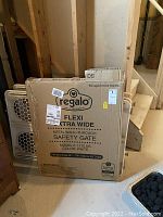 cardboard box labeled Regalo Flexi Extra Wide Metal Walk-Through Safety Gate in basement with wooden stairs and other items nearby