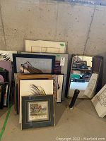 Group of framed paintings, prints, and mirror leaning against wall in basement setting showing various sizes and frame types