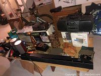 Wide shot of various items on table and floor including radios, speakers, boxed items, copper jug, badminton set, and more