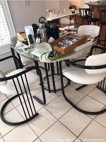 Photo showing metal and glass table with four white vinyl cushioned metal chairs. Table holds various household items not included in sale.
