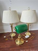 Two brass table lamps with white pleated shades and one green glass bankers lamp on a wood floor.