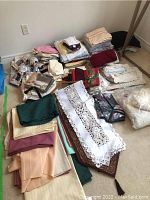 Overall view of stacks of table linens including tablecloths, runners, placemats, towels, oven mitts, and pillowcases arranged on the floor.