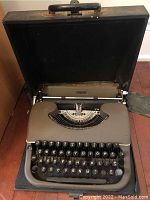 Full view of vintage metal typewriter in gray color with attached carrying case, showing keyboard and platen roller.