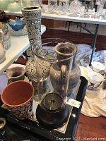 Photo showing three vases, a clear glass hurricane candle holder with a black base, and a terracotta pot with white decorative pattern.