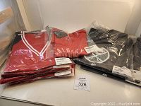 Photo showing two stacks: on left are red Calgary Flames and Detroit Red Wings shirts, on right are mostly black Buffalo Sabres shirts, all sealed in plastic with tags.