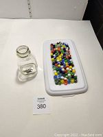 Clear glass jar with an open top, positioned next to a white tray containing multiple marbles of various colors.
