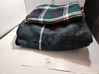 Two folded blankets stacked, green plaid on top and large black eagle blanket below, on a white background.