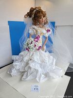 Full view of the Danbury Mint Summer Splendor bride doll standing against blue background. Doll wears white lace dress with purple flowers, holding a bouquet of white and purple flowers with a bridal veil attached.