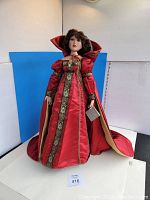Full front view of the Danbury Mint "Ruby - Winter's Princess" doll on a stand wearing a red gown with gold patterned trim and a matching hooded cape.