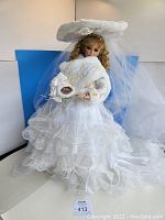 Full front view of the 26-inch Danbury Mint Winter Bliss Wedding Bride porcelain doll in white layered lace wedding dress with ruffles and holding bouquet, posed sitting with separate veil and hat seen.