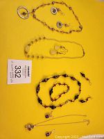 Four jewelry sets displayed against yellow backdrop, showing necklaces, earrings, and a bracelet with a lot number card.