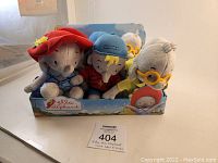 Front view of three plush toys - Ella (red hat), Frankie (blue hat), Tiki (yellow glasses) in store display box.