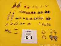 Wide top-down view of 29 pairs of varied earrings arranged on yellow background with auction lot card
