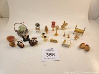 Full view of 22 miniature items laid out on white background. Includes brass miniatures of bed, candle holders, toy horse, Christmas tree, pitcher, various animal figurines, miniature bible, painted wooden clogs, raccoon figurines, bowls, and a decorative wire basket with glass bottle.