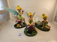 Three Tinker Bell statues on a white surface with varying poses and light features.