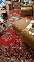 Photo showing the red machine made area rug with floral medallion patterns, beige and black accents, and fringe. Furniture and boxes partially on rug to show size and condition.