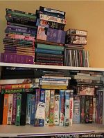 View of stacked VHS tapes, DVDs and book sets on shelves, visible titles of children's movies on VHS such as Iron Giant and Homeward Bound, various DVD cases stacked horizontally and vertically, assorted book sets hardcover and paperback.