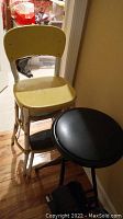 Side and angled view of the vintage metal step stool chair and collapsible stool, showing the yellow metal seat and back with rust spots and the black faux leather seat of the collapsible stool.