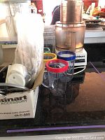 Cuisinart food processor with several clear blending cups front and center, original accessory box on left containing white discs and parts, placed on kitchen counter.