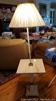 Lamp table with beige pleated lampshade, brass stand, and square marble tabletop shown from an angle on wooden floor in a living room.