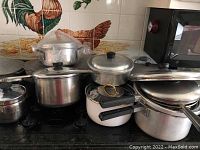Collection of various pots and pans stacked on a stovetop including Regal branded pot, some missing handles and stained from use.
