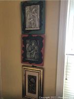 Three religious themed wall art pieces displayed on a wall. Two larger rectangular pieces in greenish-blue and reddish backings with irregular decorative edges. A smaller gold framed piece below them.