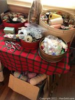 Wide view of all sewing supplies arranged on table with baskets and cardboard box underneath containing yarns. Various spools of thread, scissors, pins and notions visible.