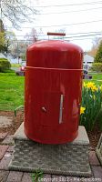Full view of cylindrical red smoker with lid and side handles on a stone platform